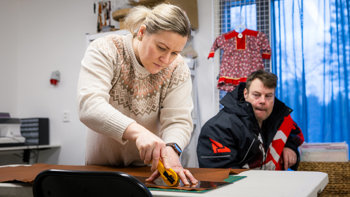 Kuvassa nainen leikkaa pöydällä poronnahkaa. Taustalla mies seuraa työtä. 