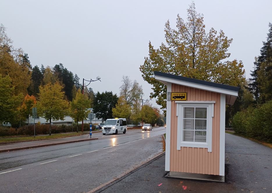 Vaaleanpunaiseksi maalattu pysäkkikatos, ohitse menee kaksi autoa. 